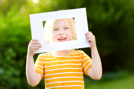 Cute cheerful little girl holding white picture frame in front of her face. Adorable child framing her smiling face.の写真素材