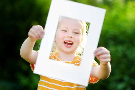 Cute cheerful little girl holding white picture frame in front of her face. Adorable child framing her smiling face.の写真素材