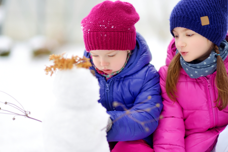 Two adorable little girls building a snowman together in beautiful winter park. Cute sisters playing in a snow. Winter activities for kids.の写真素材