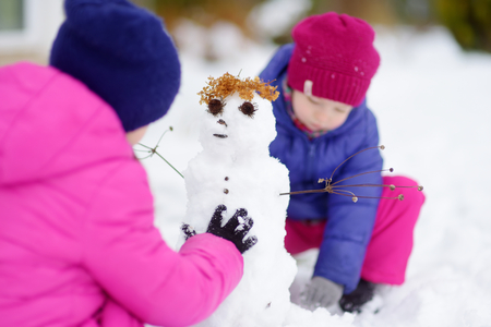 Two adorable little girls building a snowman together in beautiful winter park. Cute sisters playing in a snow. Winter activities for kids.の写真素材