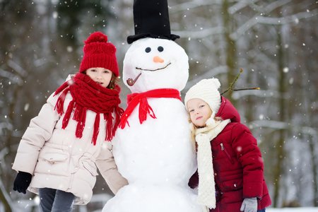 Two adorable little girls building a snowman together in beautiful winter park. Cute sisters playing in a snow. Winter activities for kids.の写真素材
