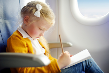 Adorable little girl traveling by an airplane. Child sitting by aircraft window and drawing a picture with colorful pencils. Traveling abroad with kids.の写真素材