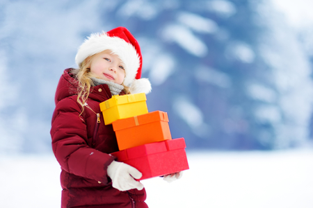 Adorable little girl wearing Santa hat holding a pile of Christmas gifts on beautiful winter day. Celebrating Xmas at home.の写真素材