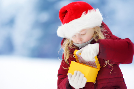 Adorable little girl wearing Santa hat holding Christmas gift on beautiful winter day. Celebrating Xmas at home.の写真素材