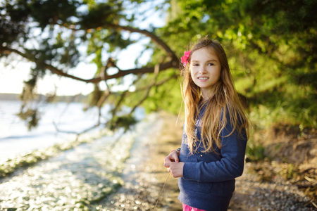 Cute little girl standing by the lake enjoying beautiful sunset view. Child exploring nature. Family activities in summer.の写真素材