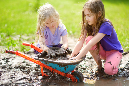 Two funny little girls playing in a large wet mud puddle on sunny summer day. Children getting dirty while digging in muddy soil. Messy games outdoors.の写真素材