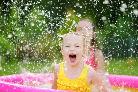 Adorable little girls playing in inflatable baby pool. Happy kids splashing in colorful garden play center on hot summer day. Summer activities for kids.の写真素材