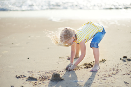 Cute little girl having fun on a sandy beach on warm and sunny summer day. Kid playing by the ocean. Summer activities for children.の写真素材