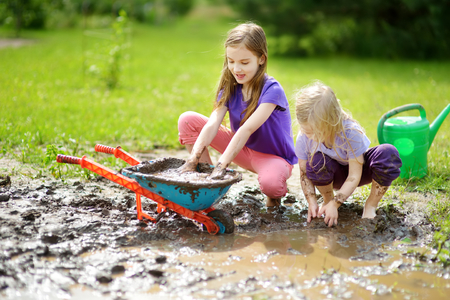 Two funny little girls playing in a large wet mud puddle on sunny summer day. Children getting dirty while digging in muddy soil. Messy games outdoors.の写真素材