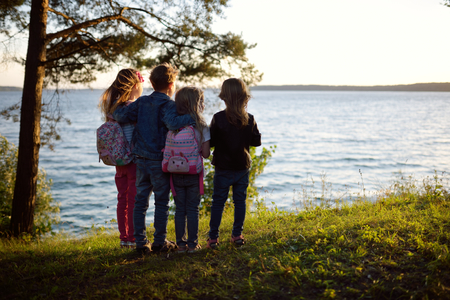 Four cute little sibling standing by the lake enjoying beautiful sunset view. Children exploring nature. Family activities in summer.の写真素材