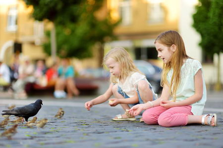 Two cute little sisters feeding birds on summer day. Children feeding pigeons and sparrows outdoors. Active leisure with kids.の写真素材