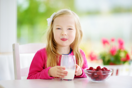 Pretty little girl eating raspberries and drinking milk at home. Cute child enjoying her healthy fresh organic fruits and berries. Healthy breakfast.の写真素材