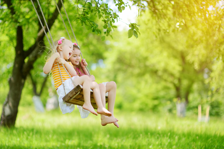 Two cute little sisters having fun on a swing together in beautiful summer garden on warm and sunny day outdoors. Active summer leisure for kids.の写真素材