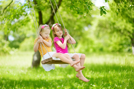 Two cute little sisters having fun on a swing together in beautiful summer garden on warm and sunny day outdoors. Active summer leisure for kids.の写真素材