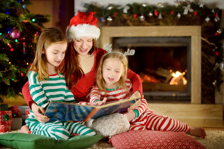 Young mother and her two little daughters reading a book by a Christmas tree in cozy living room in winterの写真素材
