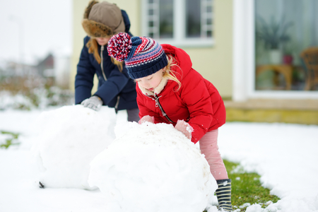 Adorable little girls building a snowman in the backyard. Cute children playing in a snow. Winter activities for kids.の写真素材