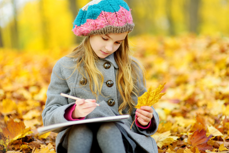 Cute little girl sketching outside on beautiful autumn day. Happy child playing in autumn park. Kid drawing with colourful pencils. Autumn activities for children.の写真素材