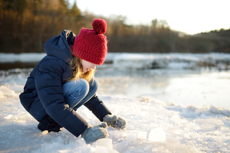 Happy little girl playing with ice blocks by frozen river during an ice break. Child having fun in winter. Winter activities for kids.の写真素材