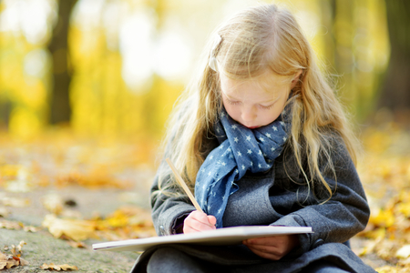Cute little girl sketching outside on beautiful autumn day. Happy child playing in autumn park. Kid drawing with colourful pencils. Autumn activities for children.の写真素材