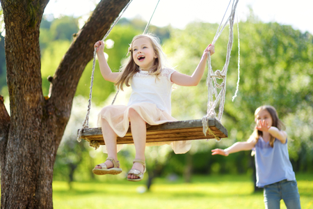 Two cute sisters having fun on a swing in blossoming old apple tree garden outdoors on sunny spring day. Spring outdoor activities for kids.の写真素材