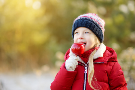 Adorable little girl eating red apple covered with sugar icing on beautiful sunny Christmas day. Child enjoying sweets, candies and gingerbread on magical Xmas time.の写真素材