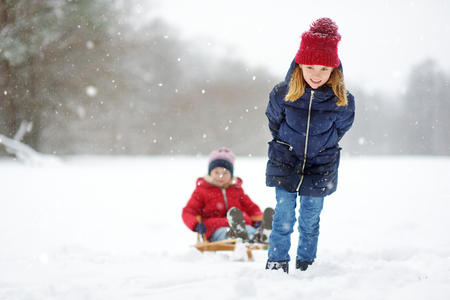 Two funny little girls having fun with a sleigh in beautiful winter park. Cute children playing in a snow. Winter activities for kids.の写真素材