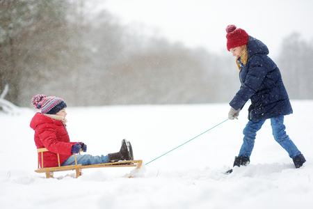 Two funny little girls having fun with a sleigh in beautiful winter park. Cute children playing in a snow. Winter activities for kids.の写真素材