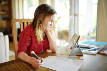Smart preteen schoolgirl doing her homework with digital tablet at home. Child using gadgets to study. Education and learning for kids.の写真素材