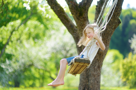 Cute little girl having fun on a swing in blossoming old apple tree garden outdoors on sunny spring day. Spring outdoor activities for kids.の写真素材