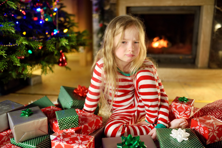 Cute little girl feeling unhappy with her Christmas gifts. Child sitting by a fireplace in a cozy dark living room on Xmas eve. Too many presents for Christmas.の写真素材