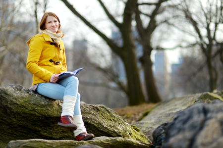 Happy young woman tourist reading a book at Central Park in New York City. Female traveler enjoying views of downtown Manhattan. Travelling in USA.の写真素材