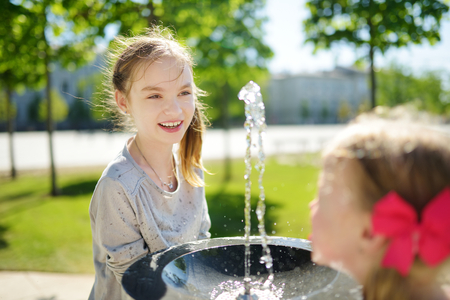 Two sisters having fun with drinking water fountain on warm and sunny summer day. Kids playing outdoors.の写真素材