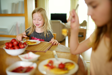 Cute little sisters enjoying their breakfast at home. Pretty children eating pancakes with strawberry sauce before school. Healthy nutrition for children.の写真素材