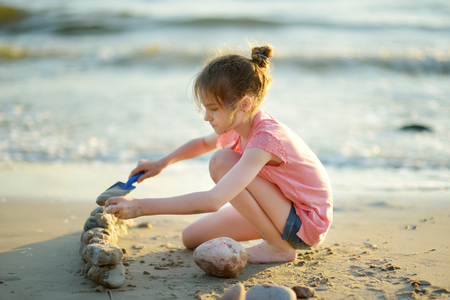Cute little girl having fun on a sandy beach on warm and sunny summer day by the sea. Kid playing by the ocean. Summer activities for children.の写真素材