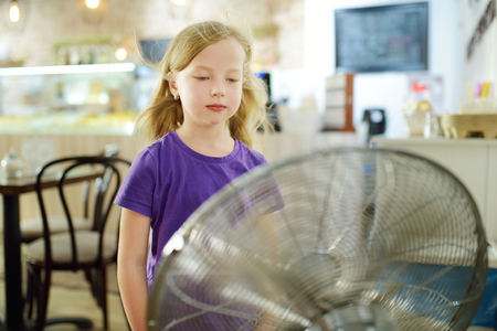 Cute little girl standing in front of a fan on hot summer day. Child enjoying cool wind in summer season. Hot weather conditions.の写真素材