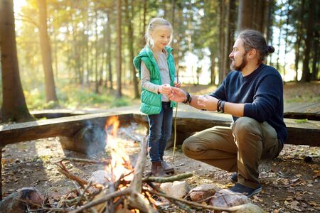 Cute young girl learning to start a bonfire. Father teaching her daughter to make a fire. Child having fun at camp fire. Camping with kids in fall forest. Family leisure with kids at autumn.の写真素材