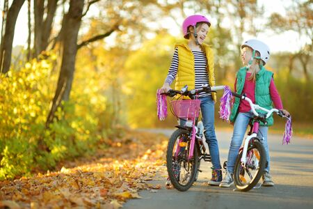 Cute little sisters riding bikes in a city park on sunny autumn day. Active family leisure with kids. Children wearing safety helmet while riding a bicycle.の写真素材