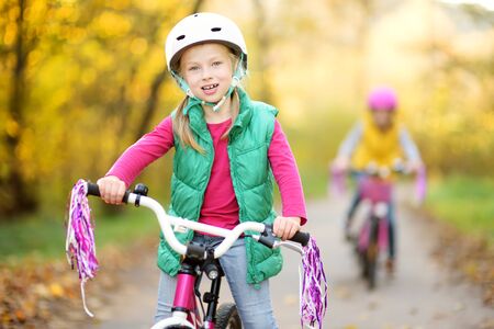 Cute little sisters riding bikes in a city park on sunny autumn day. Active family leisure with kids. Children wearing safety helmet while riding a bicycle.の写真素材