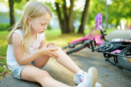 Cute little girl sitting on the ground after falling off her bike at summer park. Child getting hurt while riding a bicycle. Active family leisure with kids.の写真素材