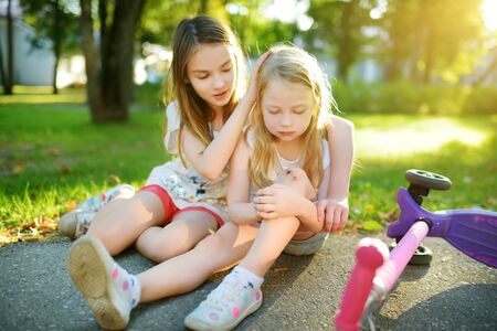 Adorable girl comforting her little sister after she fell off her scooter at summer park. Child getting hurt while riding a kick scooter. Active family leisure with kids.の写真素材