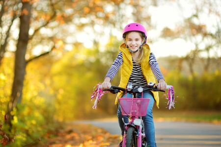 Adorable young girl riding a bike in a city park on sunny autumn day. Active family leisure with kids. Child wearing safety helmet while riding a bicycle.の写真素材