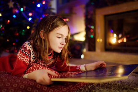 Happy young girl reading a story book by a fireplace in a cozy dark living room on Christmas eve. Celebrating Xmas at home. Winter evening with family and kids.の写真素材