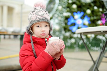 Cute young girl drinking hot chocolate on traditional Christmas fair in Vilnius, Lithuania. Child enjoying sweets, candies and gingerbread on Xmas market. Winter time with family and kids.の写真素材