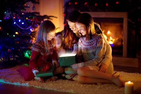 Happy young mother and her two small daughters opening a magical Christmas gift by a fireplace in a cozy dark living room on Christmas eve. Winter evening at home with family and kids.の写真素材