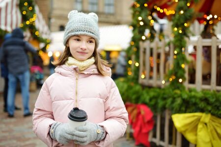 Cute young girl drinking hot chocolate on traditional Christmas fair in Riga, Latvia. Child enjoying sweets, candies and gingerbread on Xmas market. Winter time with family and kids.の写真素材
