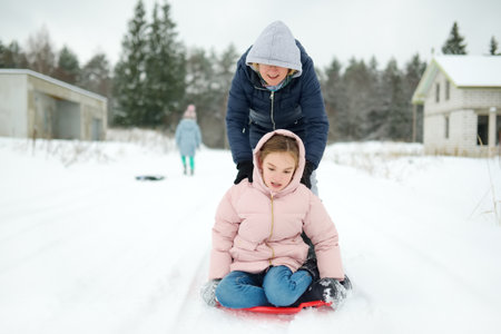 Funny young girl and her grandma having fun with a sleigh in beautiful winter park. Cute child playing in a snow. Winter activities for kids.の写真素材
