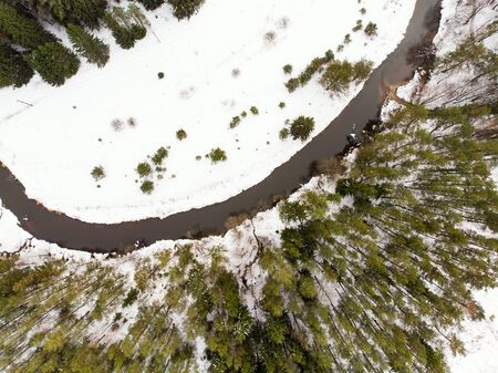 Aerial top down view of narrow river flowing between snow covered trees. Chilly winter day. Scenic...