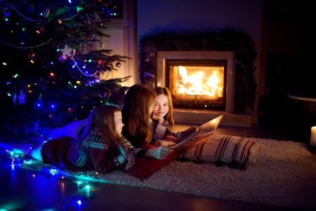Happy young mother and her daughters reading a story book together by a fireplace in a cozy dark living room on Christmas eve. Celebrating Xmas at home. Winter evening with family and kids.の写真素材