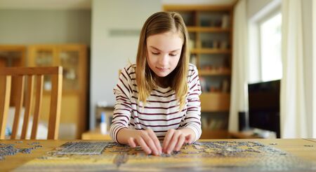 Cute young girl playing puzzles at home. Child connecting jigsaw puzzle pieces in a living room table. Kid assembling a jigsaw puzzle. Fun family leisure. Stay at home activity for kids.の写真素材