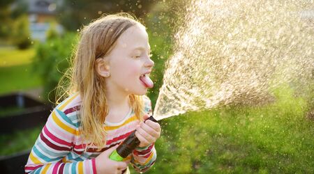 Adorable little girl playing with a garden hose on warm summer day. Child having fun with water on...